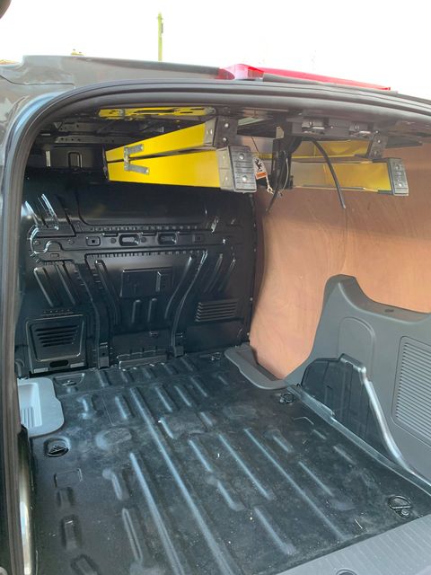 The open rear cargo area of a vehicle showing a black textured floor mat, black molded side panels, and a tan interior lining, with a yellow folded ladder or rack mounted near the ceiling.